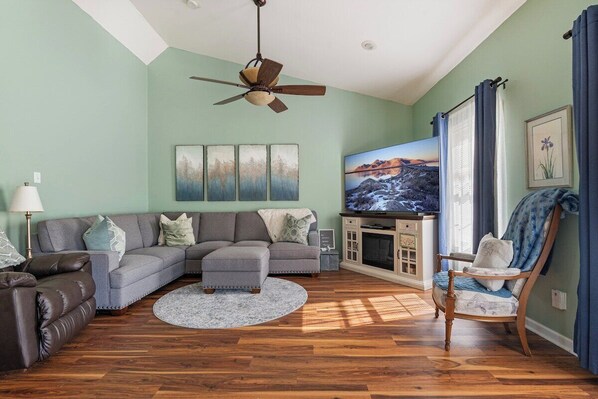 A living room with a sectional sofa, a wooden floor, a ceiling fan, a television, and a fireplace.