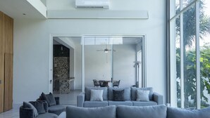 A modern living room with a grey sectional sofa, a glass door leading to a dining area, and a large window with a view of greenery. - Luxurious Getaway w Private Pool Garden (Santiago)