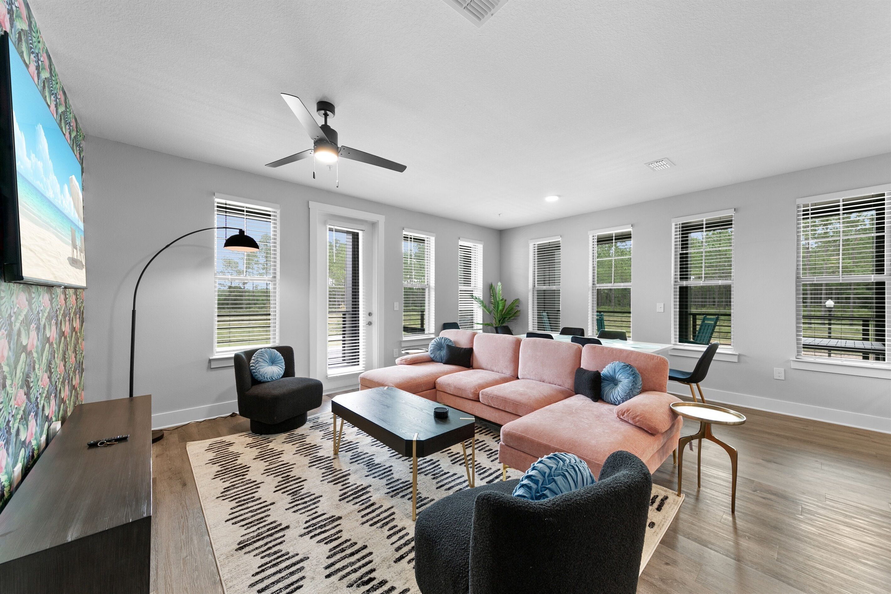 A spacious living room with a large sofa, a coffee table, and a TV mounted on the wall. There are multiple windows with blinds, a ceiling fan, and a floor lamp.