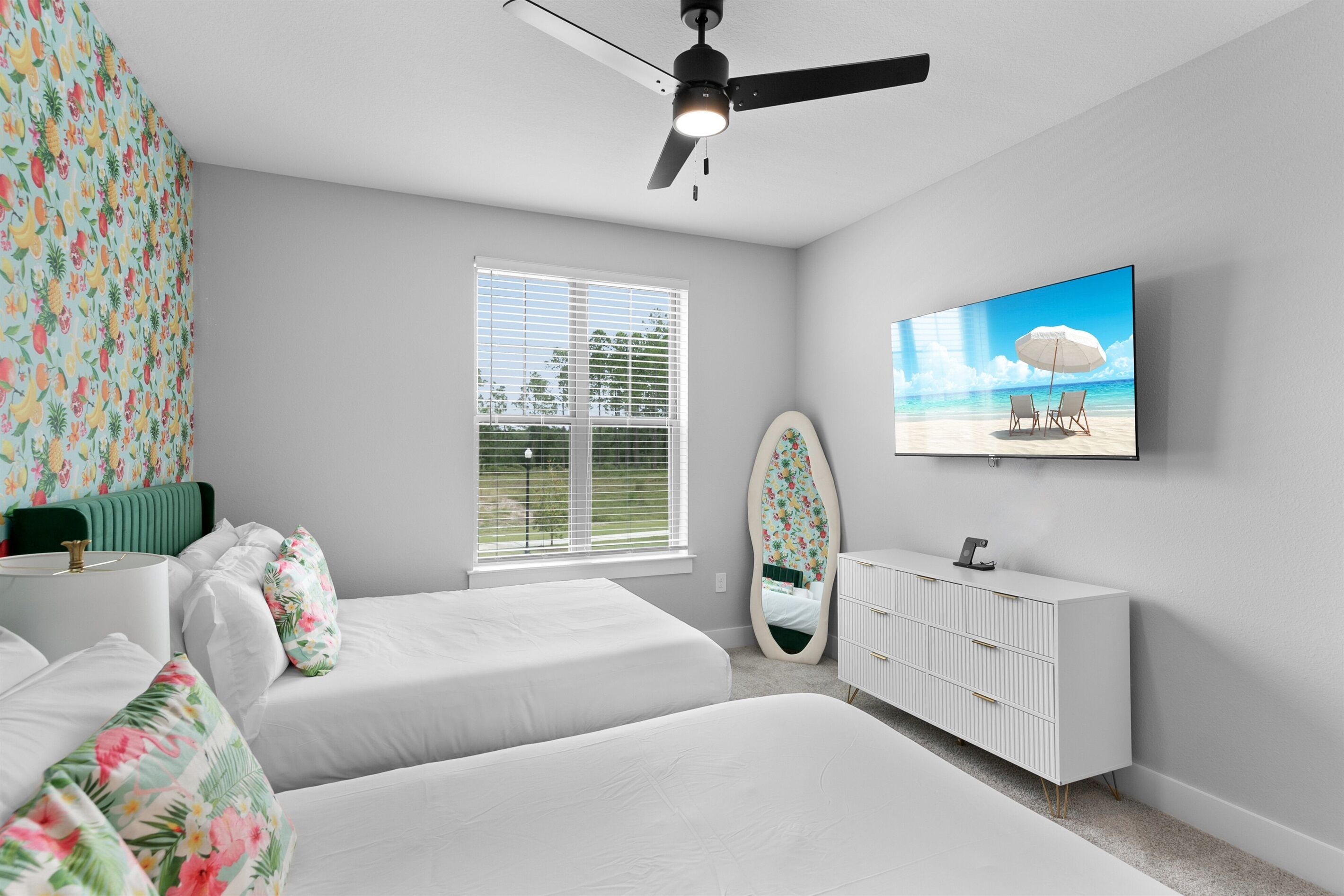 A bedroom with two beds, a vanity, a ceiling fan, a window with blinds, and a TV on the wall.
