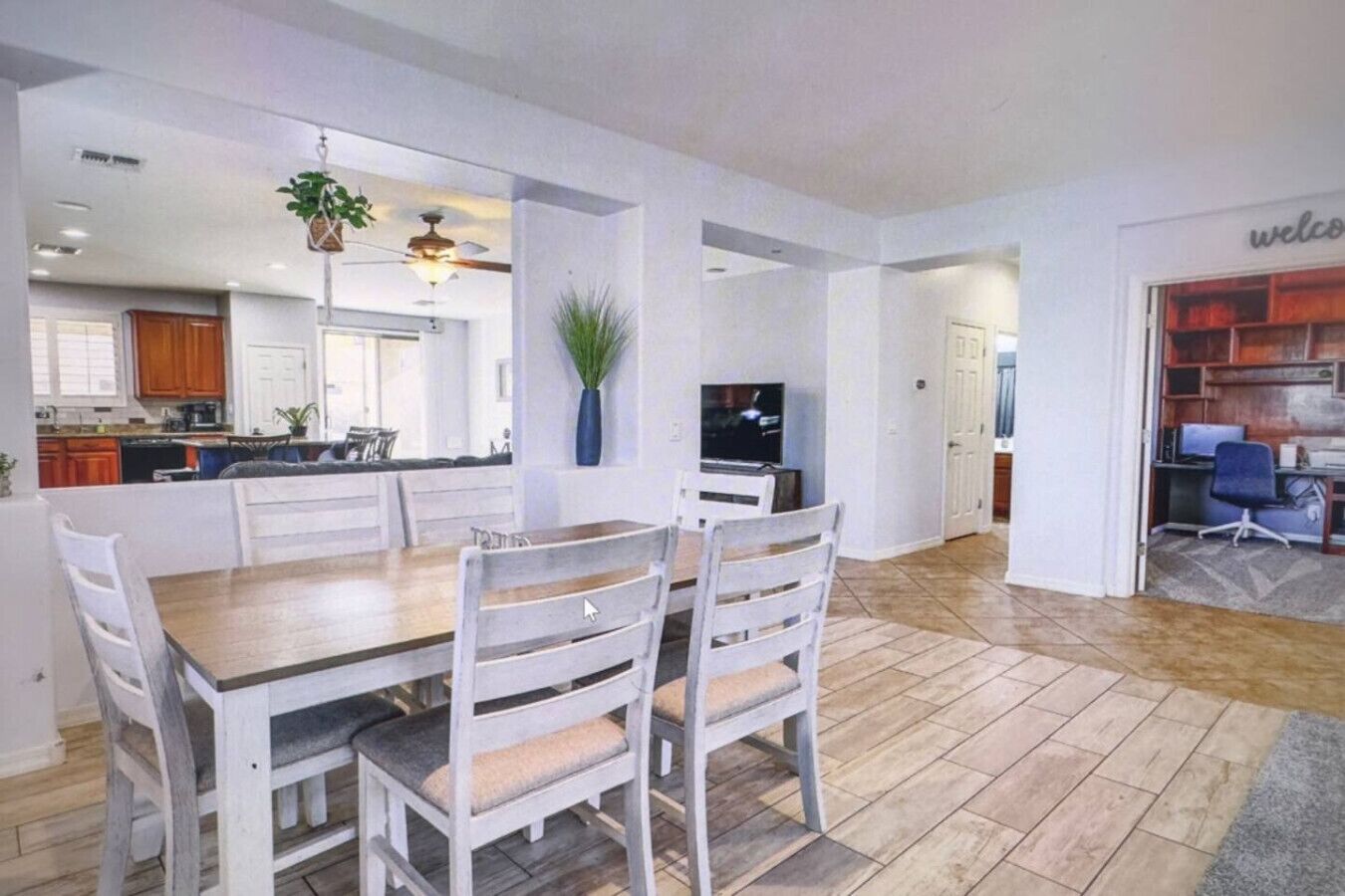 A spacious dining area with a wooden table and white chairs, a kitchen in the background, and a living area with a TV and seating.