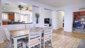 Un comedor amplio con una mesa de madera y sillas blancas, una cocina al fondo y una sala de estar con televisor y asientos.