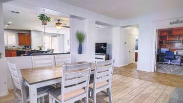 A spacious dining area with a wooden table and white chairs, a kitchen in the background, and a living area with a TV and seating.