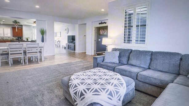 Un salón espacioso con un sofá gris, un otomano estampado y una zona de comedor con sillas de madera y una mesa.