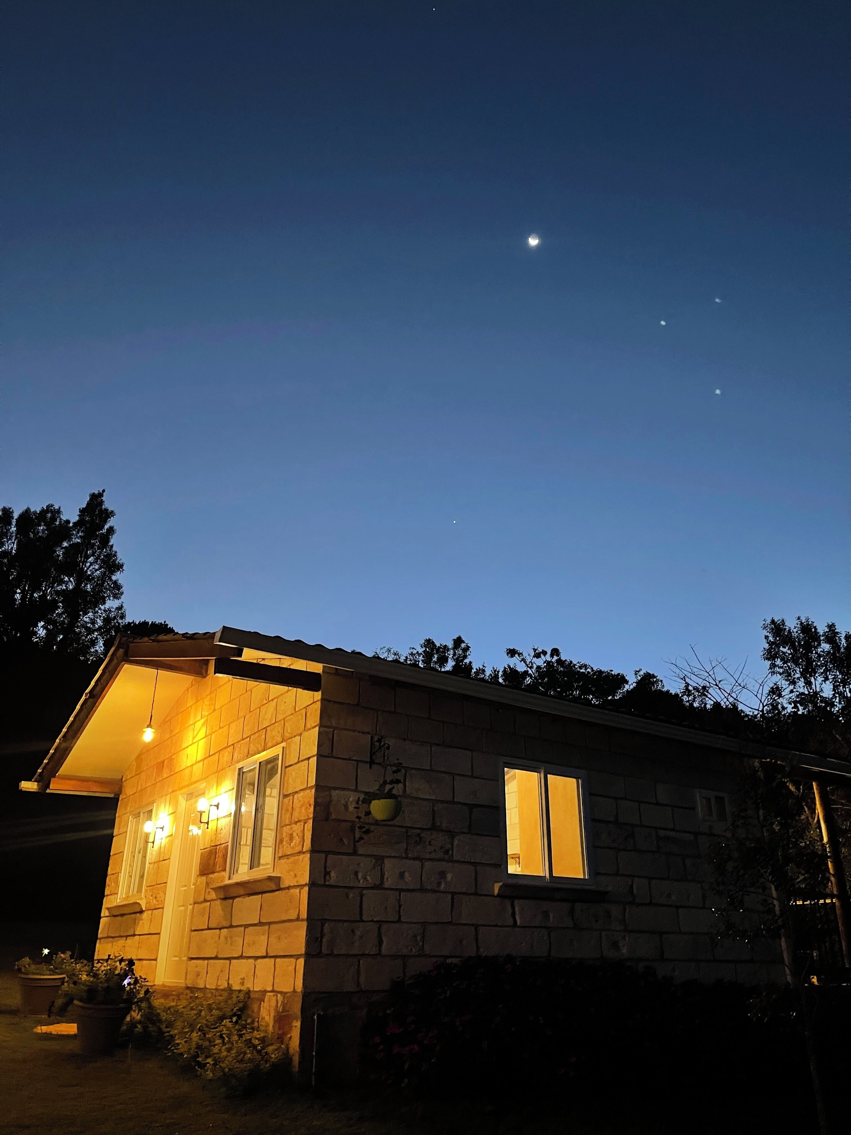 Une maison avec une façade en briques, un porche orné de plantes en pot et un ciel nocturne dégagé où l’on peut voir les étoiles.
