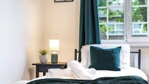 A neatly made bed with a dark green throw, a bedside lamp, and a small potted plant on a nightstand.