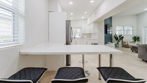 A modern kitchen with white cabinets, a white countertop, and a dining area with a white table and black chairs. - 5BD Beach House, Sun, Pool, BBQ & Shop! Beach & Aventura Mall – Sleeps 26! (Miami)
