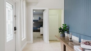 A hallway with a door leading to a bedroom, a console table with a plant, and a door with a blue panel. - 5BD Beach House, Sun, Pool, BBQ & Shop! Beach & Aventura Mall – Sleeps 26! (Miami)