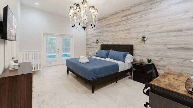 A bedroom with a bed, a desk, a chair, a chandelier, and a wooden wall. - 5BD Beach House, Sun, Pool, BBQ & Shop! Beach & Aventura Mall – Sleeps 26! (Miami)
