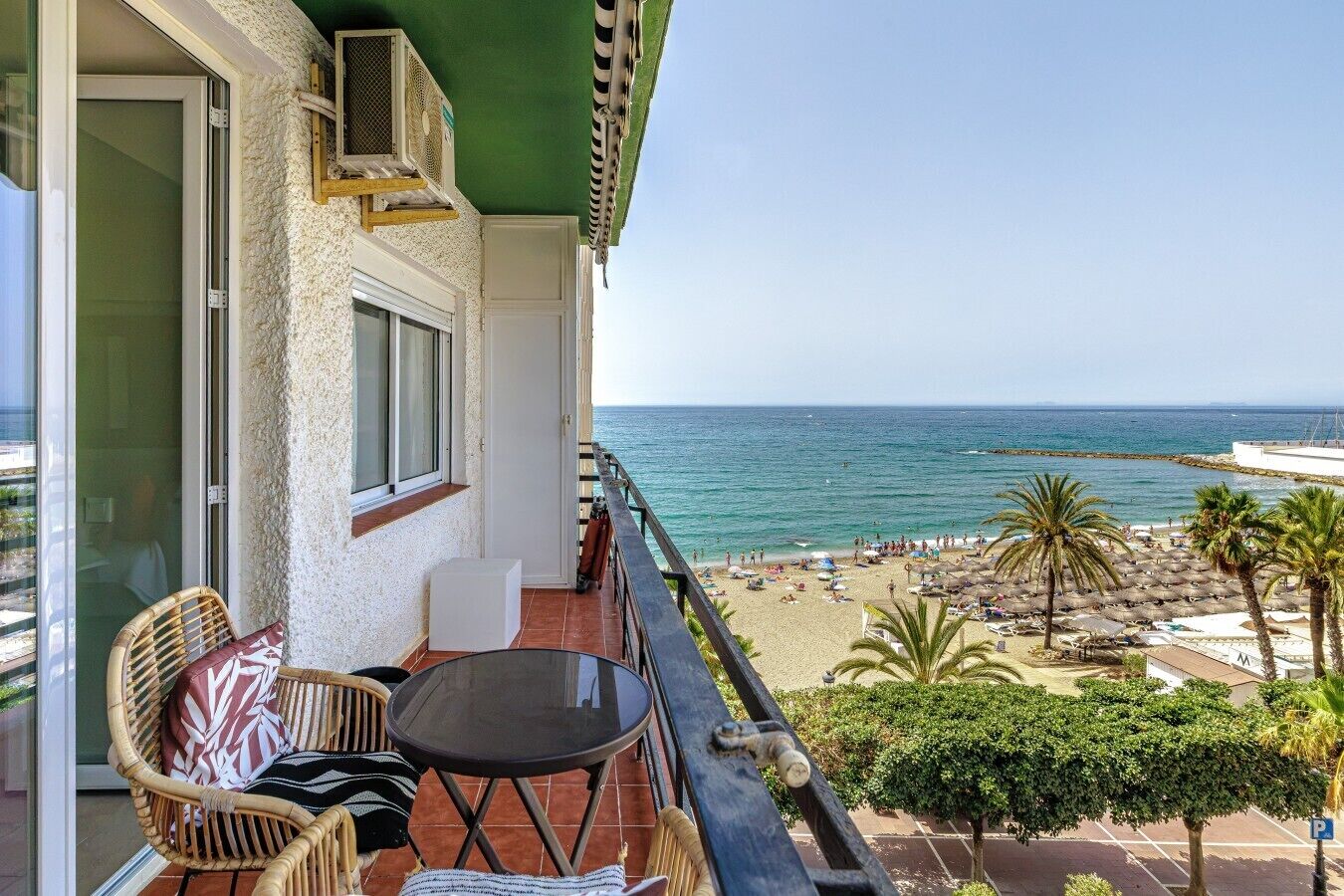 A balcony with a table and chairs, overlooking a beach with umbrellas and palm trees.