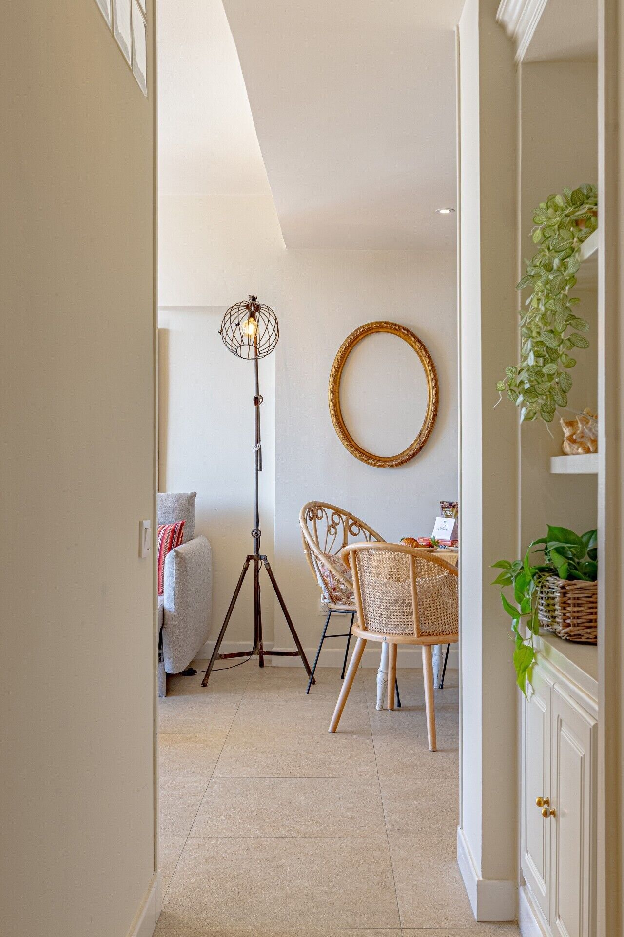 A hallway with a floor lamp, two wicker chairs, a round mirror, and a cabinet with a plant.