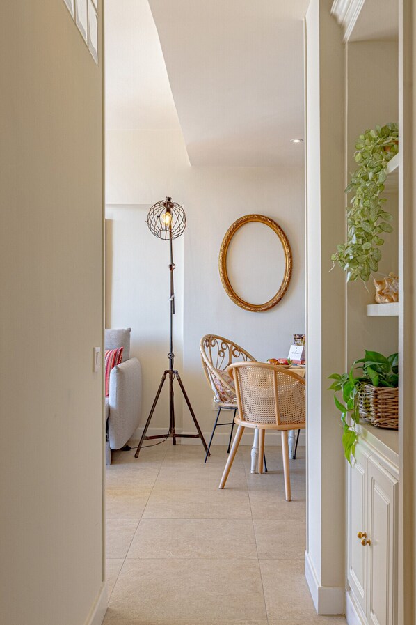 Un couloir avec un lampadaire, deux fauteuils en osier, un miroir rond et un meuble orné de plantes.