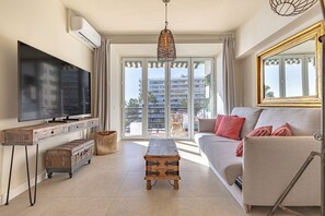 A living room with a flat-screen TV, a wooden coffee table, a sofa with cushions, a wicker hanging light, and a large window with a view of buildings.