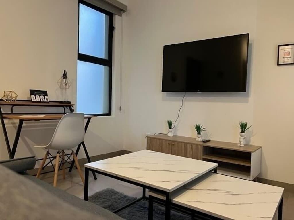 A modern living room with a flat-screen TV mounted on the wall, a wooden desk with a chair, and a sofa.