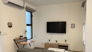 A modern living room with a grey sofa, a flat-screen TV mounted on the wall, a wooden side table, and a small cabinet with decorative plants. - Cozy 2-bedroom condo with rooftop terrace w/arch view in Cabo San Lucas (cabo san lucas)