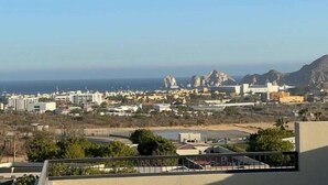 A coastal cityscape with buildings, a clear sky, and mountains in the distance. - Cozy 2-bedroom condo with rooftop terrace w/arch view in Cabo San Lucas (cabo san lucas)