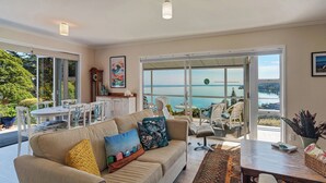 A spacious living room with a large window overlooking a body of water, a dining area with a table and chairs, and a cozy seating area with a sofa and cushions.