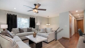 A modern living room with a sectional sofa, a coffee table, a floor lamp, and a ceiling fan. - Modern 4BR Gretna NE, Garage, Fenced Yard, W/D, Pet-Friendly, Near Outlets (Gretna)