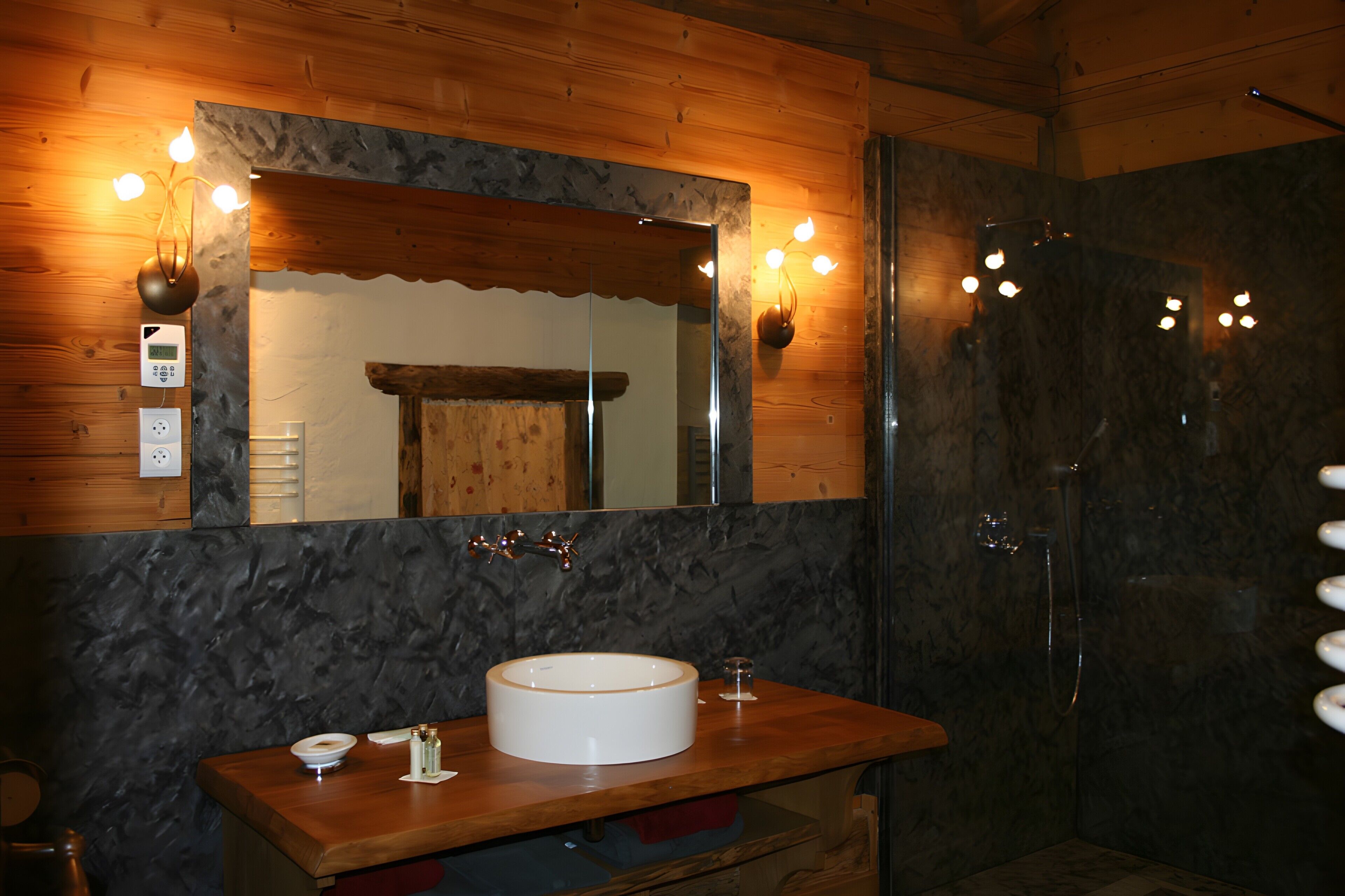 Un baño con tocador de madera, un espejo grande, un lavamanos redondo, una ducha y una lámpara de pared.