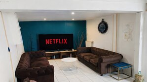 A living room with two brown sofas, a white coffee table, a television with a Netflix logo, a clock, and a map on the wall. - Gîte Le Flot House with Private Garden and Wi-Fi (Saint-Florentin)