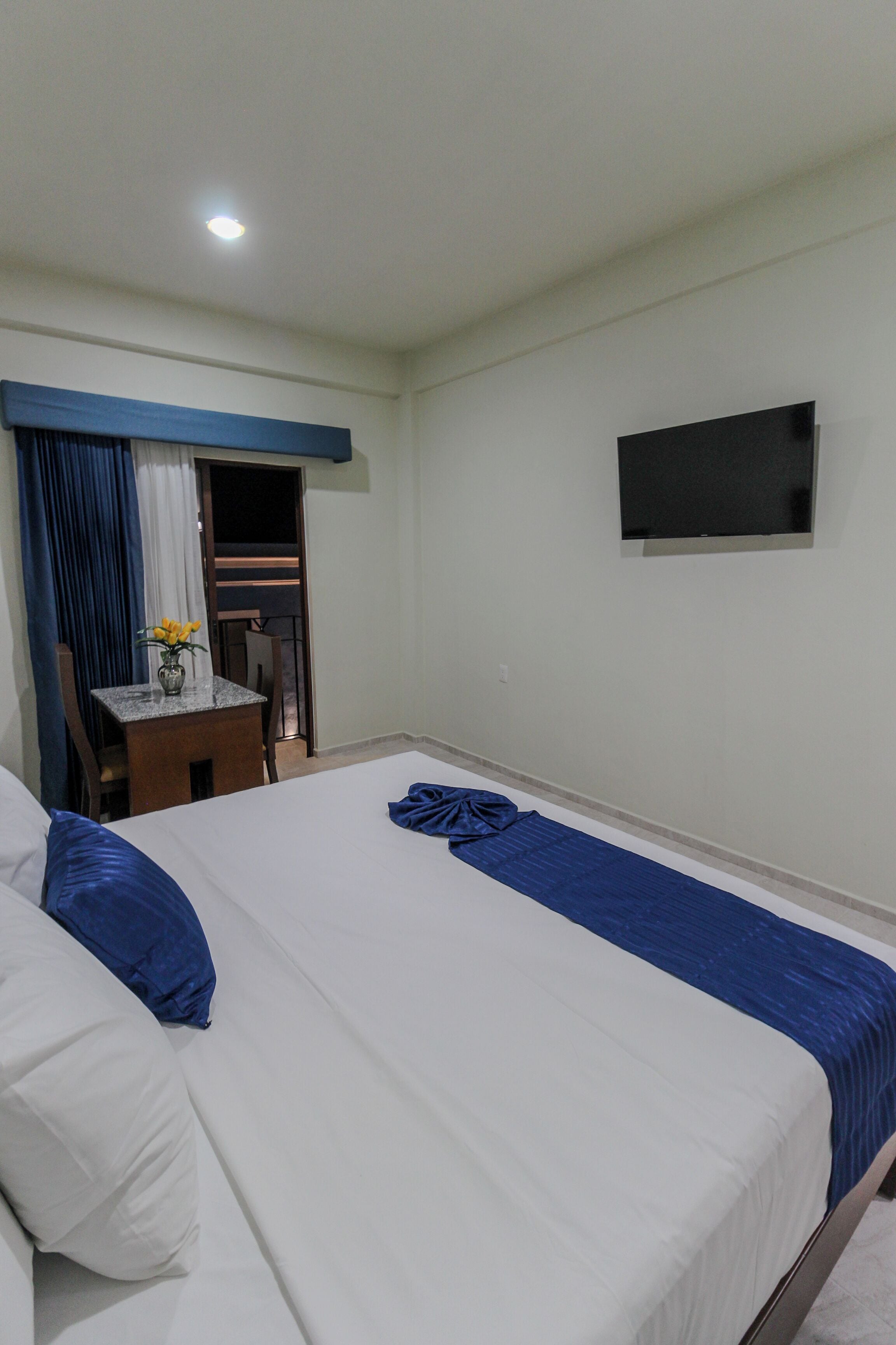 A hotel room with two beds, a small table with a vase of flowers, a TV on the wall, and a balcony with a chair.