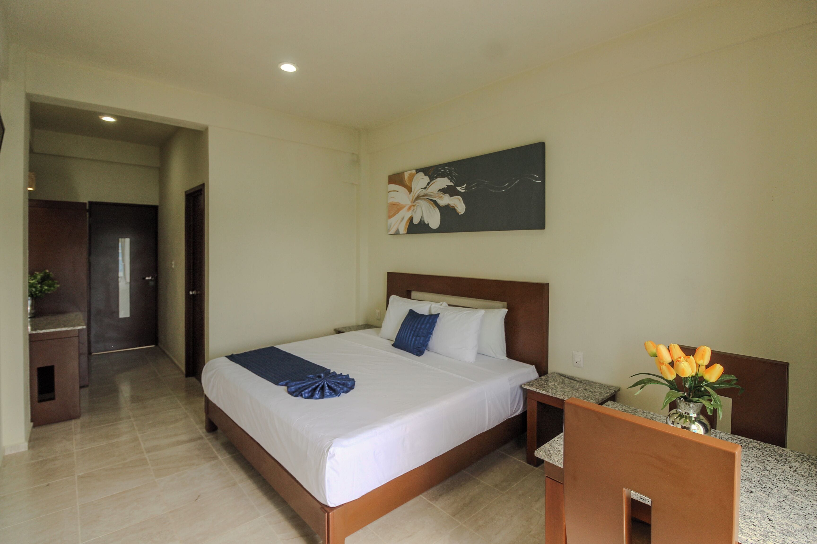 A hotel room with a bed, a nightstand, a vase with flowers, and a painting on the wall.
