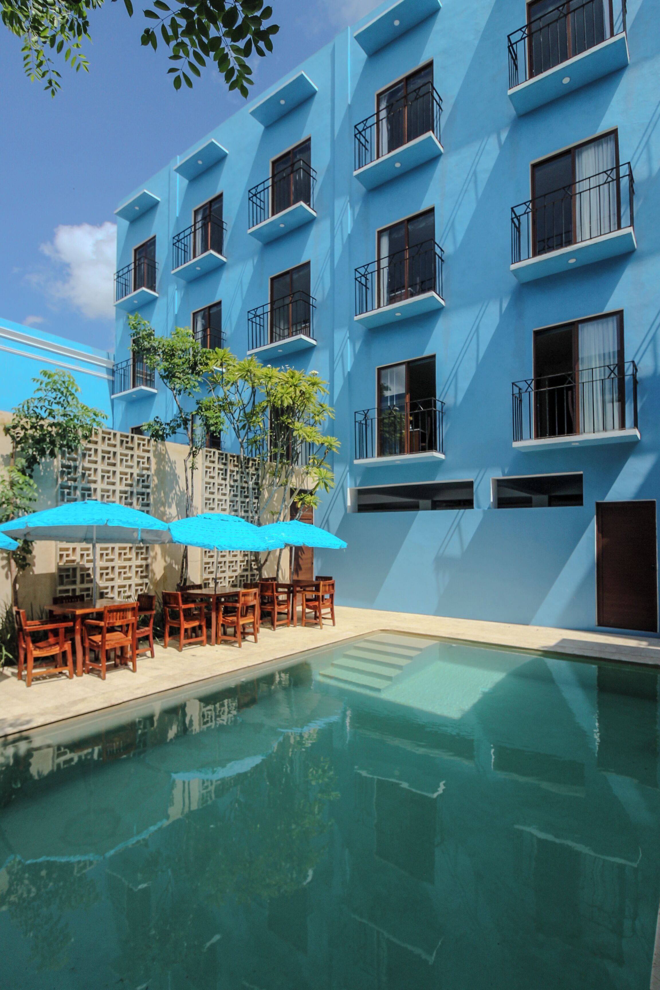 Un bâtiment bleu doté de balcons, d’une piscine et de tables en bois surmontées de parasols bleus.