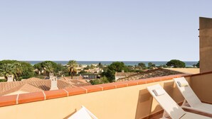 A balcony with two white lounge chairs, a view of the sea, and a townscape with buildings and trees.