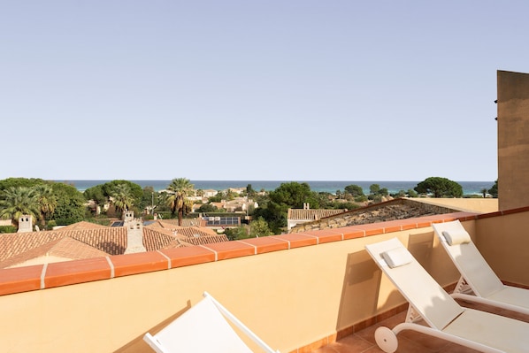 A balcony with two white lounge chairs, a view of the sea, and a townscape with buildings and trees.