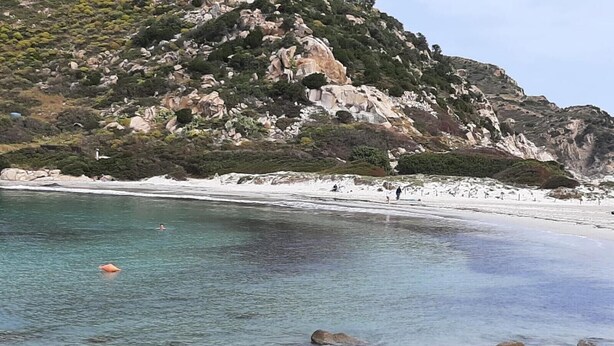 A rocky beach with clear water, a sandy shore, and a mountainous backdrop.