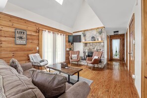 A cozy living room with a wooden ceiling, a stone fireplace, and a television mounted on the wall.