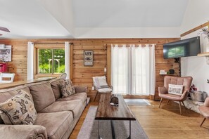 A cozy cabin-style living room with a wooden interior, a large window with curtains, a flat-screen TV mounted on the wall, and a comfortable seating area with a sofa and armchairs.