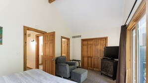 Un dormitorio con una cama, un sillón gris, un pequeño reposapiés, un televisor y armarios de madera.