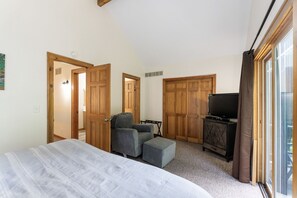 Une chambre à coucher avec un lit, un fauteuil gris, un petit pouf, une télévision et des armoires en bois.