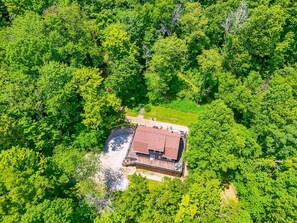 An aerial view of a property surrounded by dense green foliage, featuring a large building with a red roof, a swimming pool, and a wooden deck.