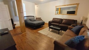 Una sala de estar con un sofá de cuero marrón, un sofá gris, una mesa de centro, suelo de madera, una puerta blanca y un espejo en la pared.