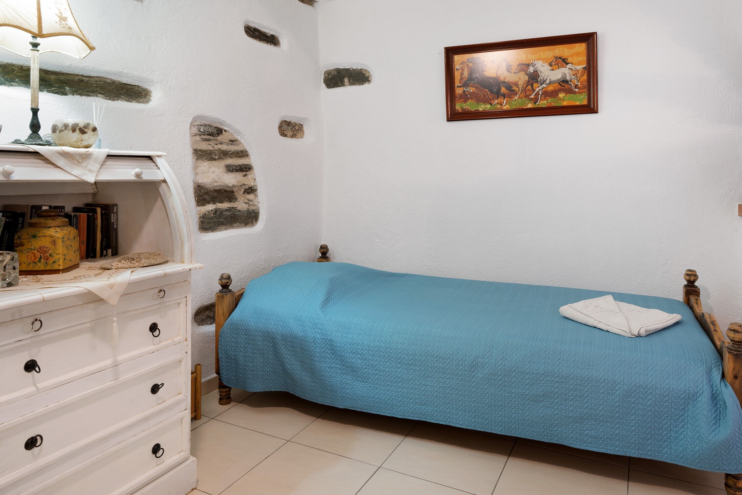 A bedroom with a bed, a dresser, a lamp, a painting, and a tiled floor.