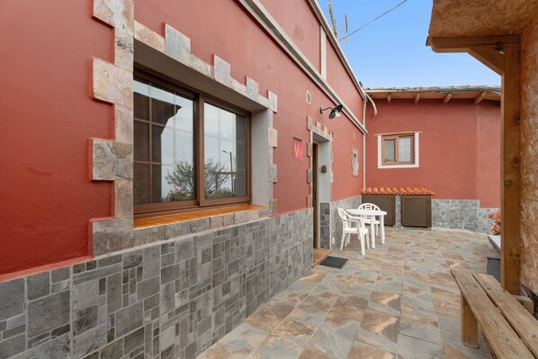 Ein Gebäude mit roter Fassade, einer Steinwand, einem Fenster, einer Tür und einer kleinen Terrasse mit einer Bank und einem Tisch.