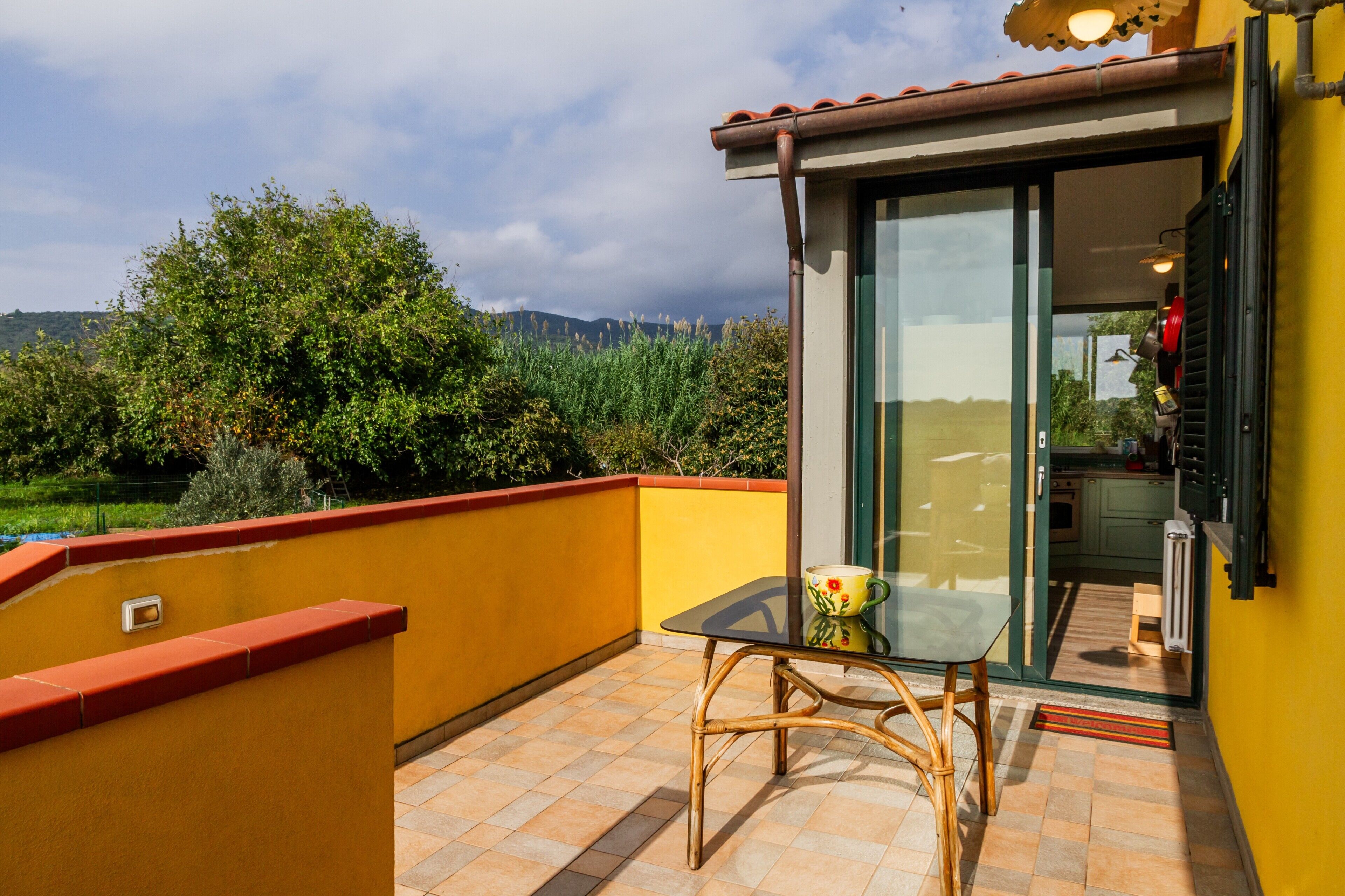 Un balcon avec une table et des chaises, une porte vitrée donnant sur une cuisine, et une vue sur la verdure et les collines.