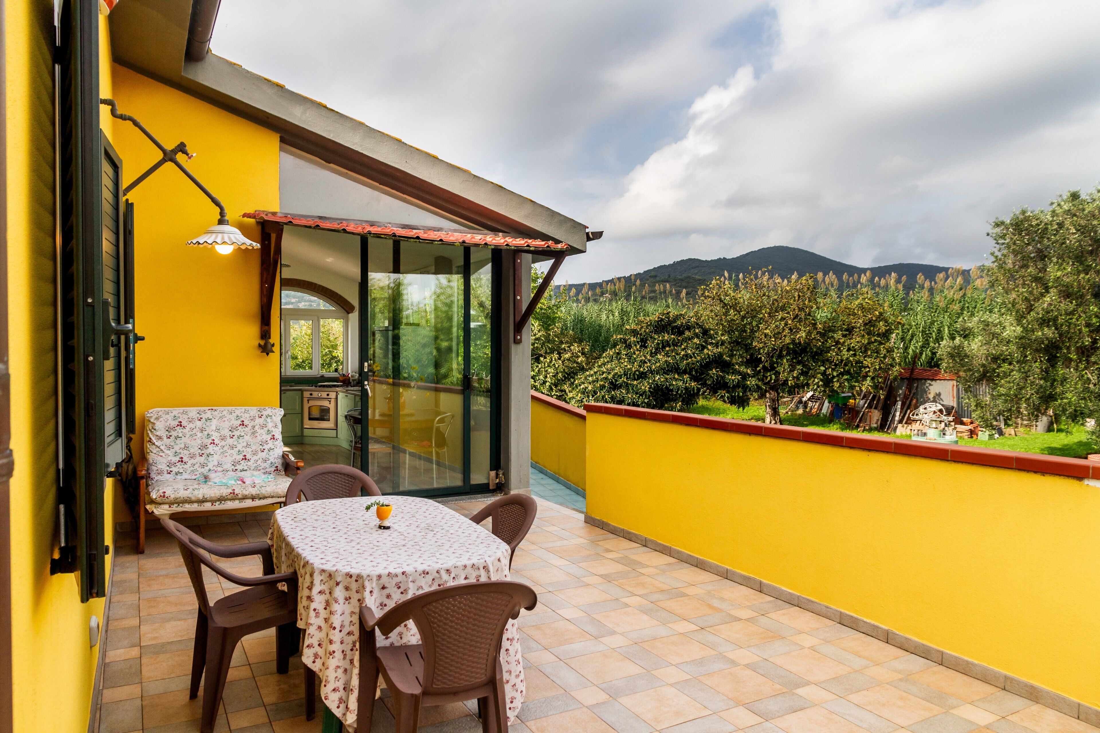 Une terrasse extérieure avec une table et des chaises, une porte vitrée donnant sur une cuisine, et une vue sur un jardin et des montagnes.