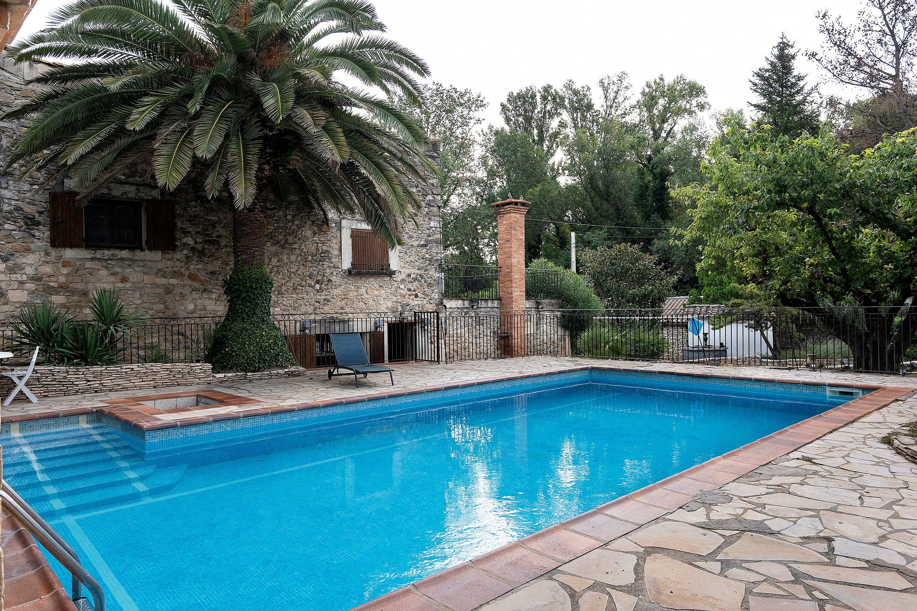 Ein Pool mit Steinfliesen, eine Palme und ein Steinhaus mit einem Fenster.