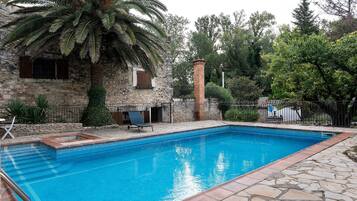 Ein Pool mit Steinfliesen, eine Palme und ein Steinhaus mit einem Fenster.