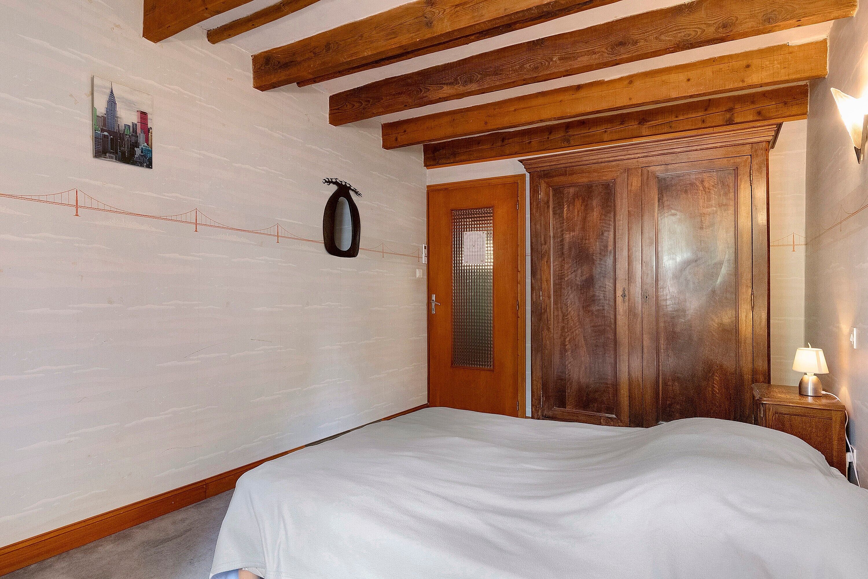 Een slaapkamer met een bed, houten balken, een deur met raam en een spiegel aan de muur.