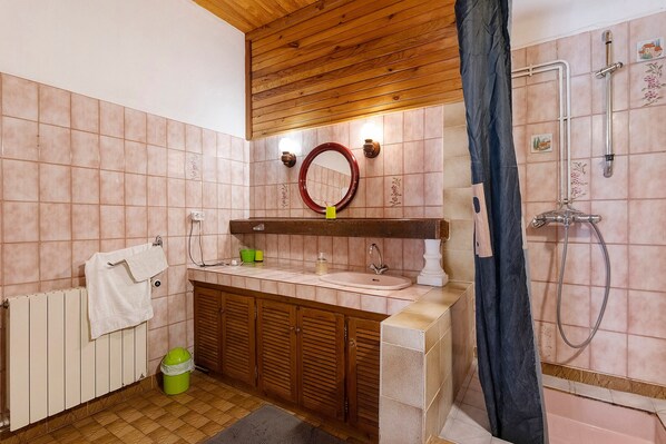 A bathroom with a wooden ceiling, a sink with a mirror above it, a shower with a hose, and a radiator.
