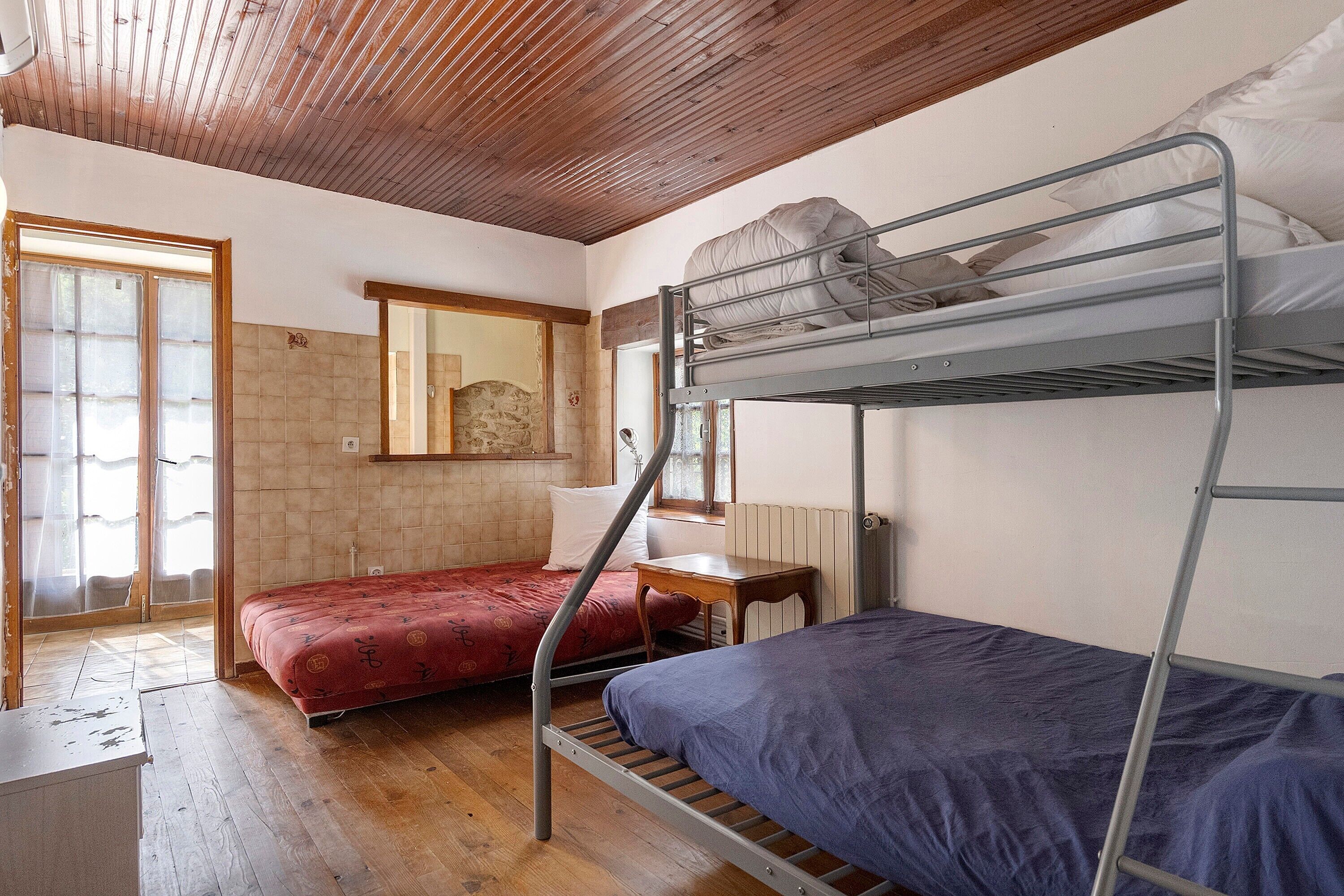 A dormitory room with bunk beds, a wooden ceiling, a window with blinds, and a tiled wall.