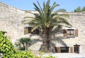 Un bâtiment en pierre avec un palmier devant, des volets en bois et un portail en métal.