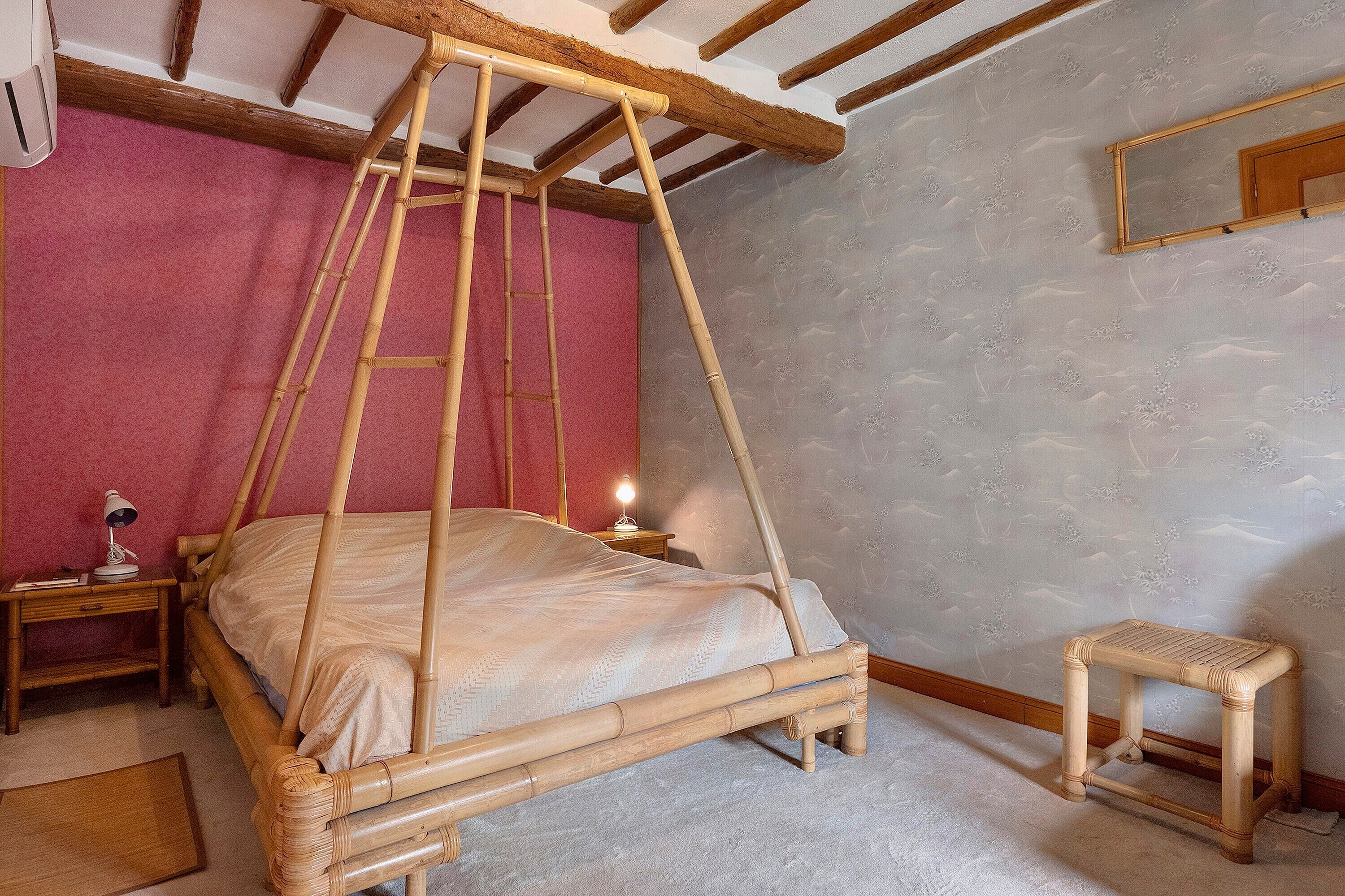 A room with a four-poster bed, a nightstand with a lamp, a wooden stool, and a mirror on the wall.