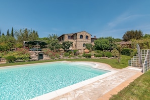 Una piscina con cubierta de piedra, una casa de piedra con ventanales arqueados y una pérgola verde.