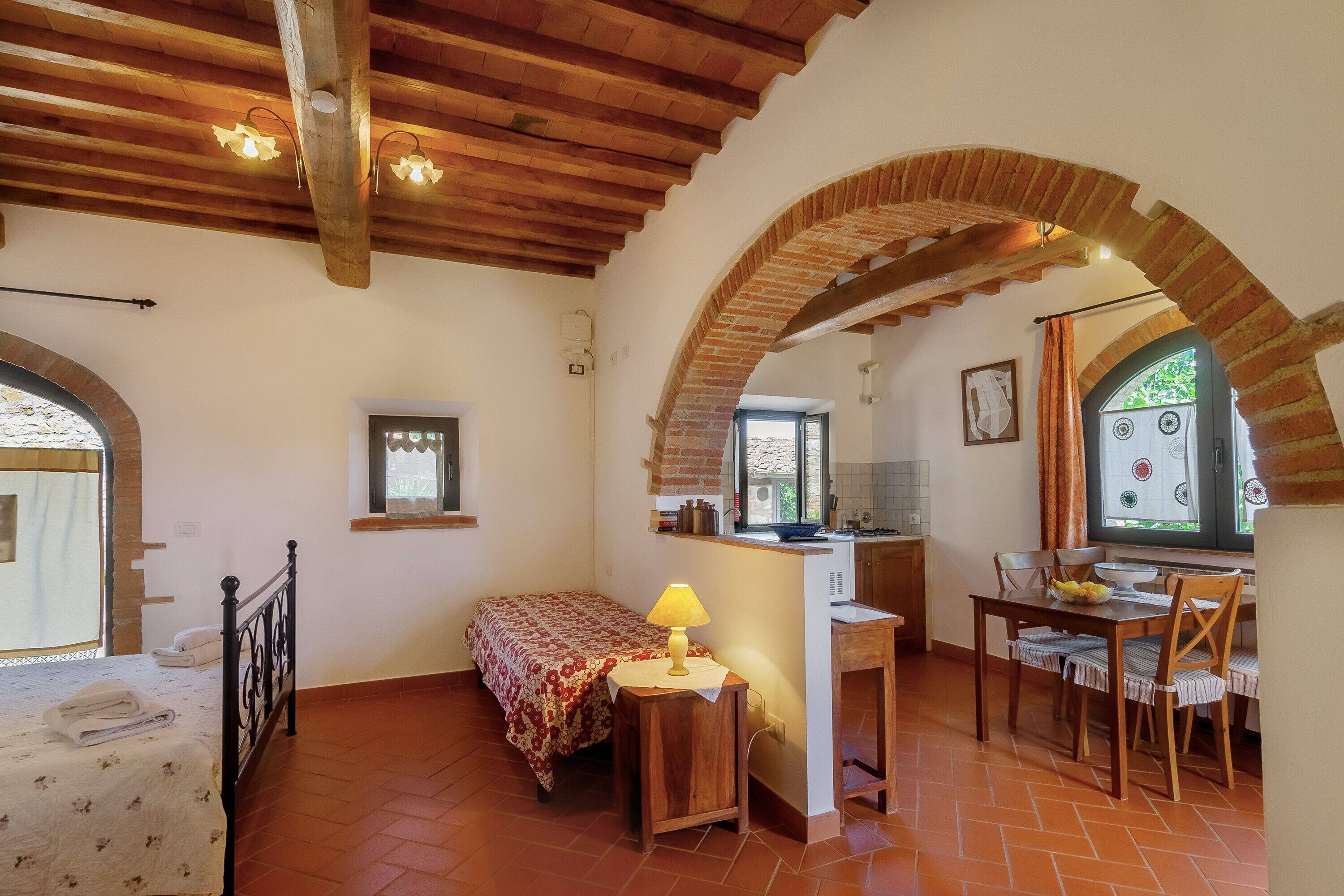 A cozy room with a bed, a desk with a lamp, a dining table with chairs, and a kitchen area.