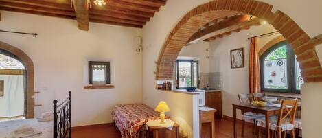 A cozy room with a bed, a desk with a lamp, a dining table with chairs, and a kitchen area.